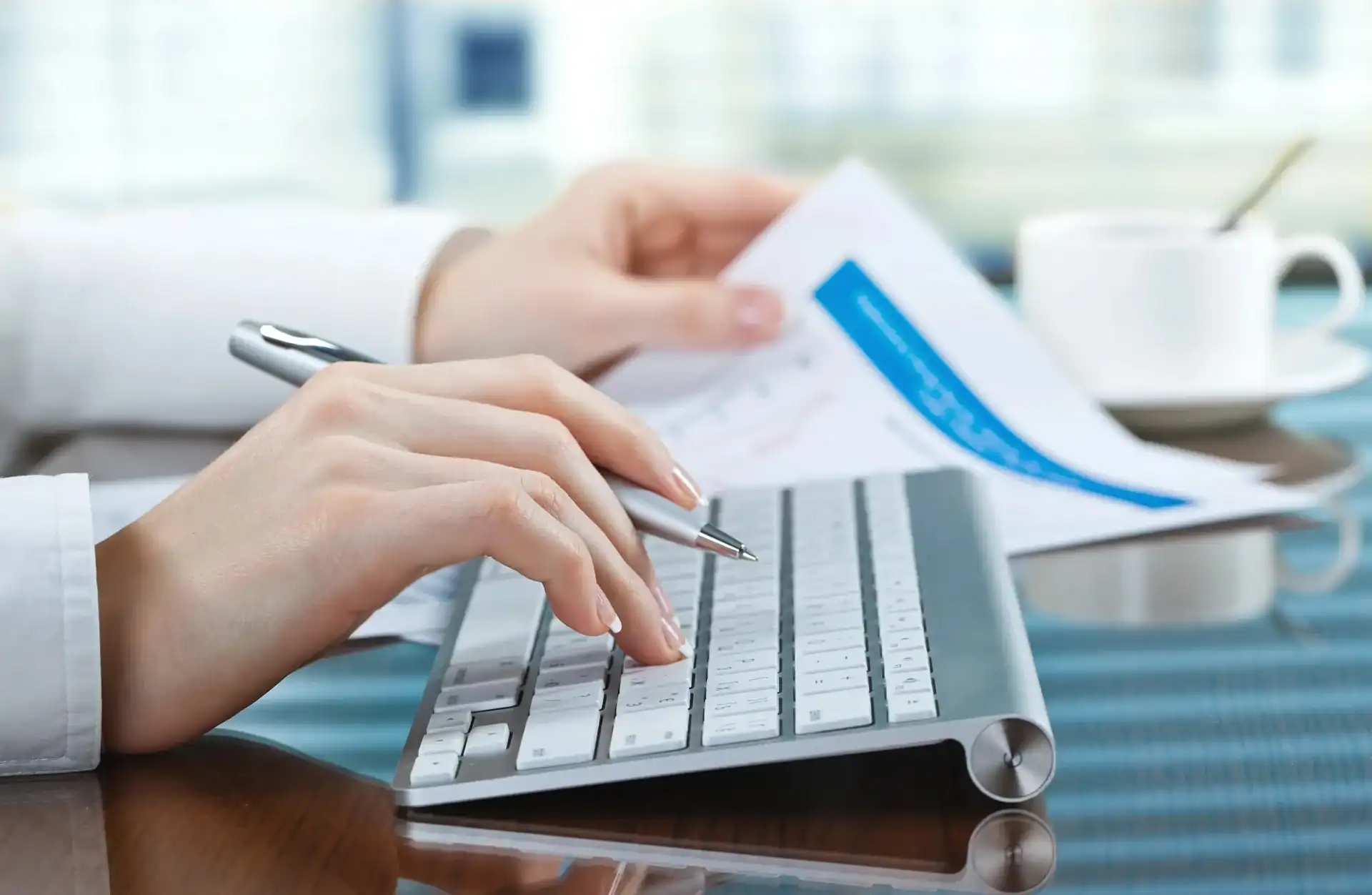 Person using a computer keyboard with one hand while holding a document and pen with the other; a white coffee cup is on the desk in the background.