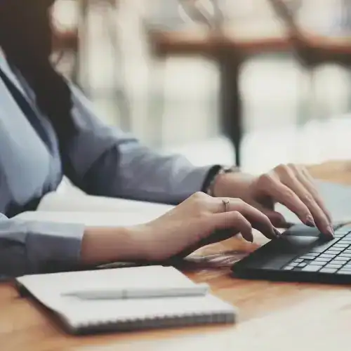Person typing on a laptop at a wooden desk with a notebook and pen beside them.