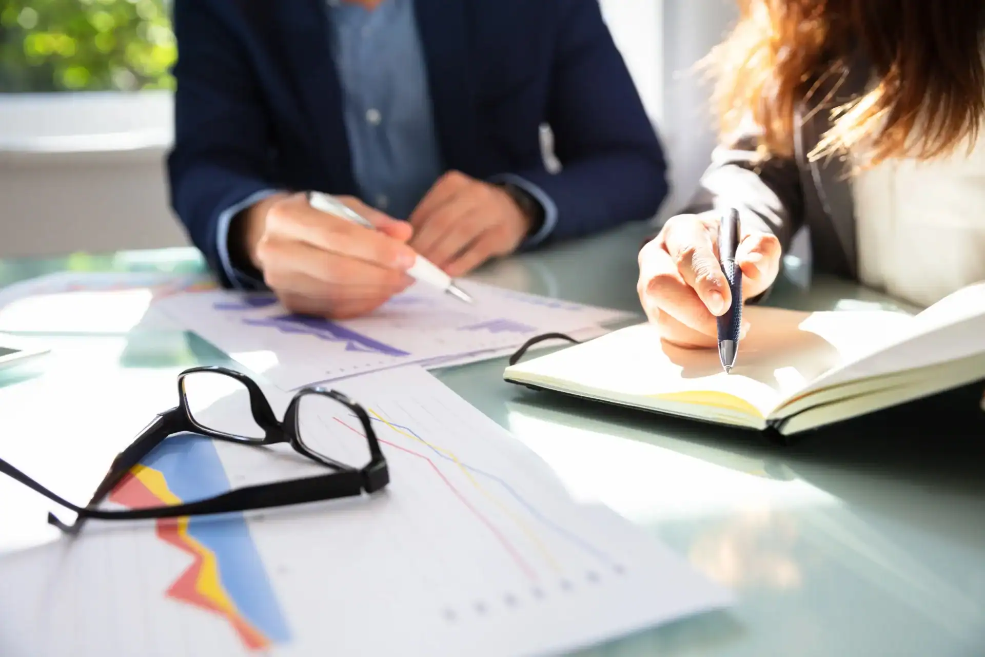 Two people in business attire review charts and make notes at a desk, with documents, a notebook, and a pair of glasses visible.